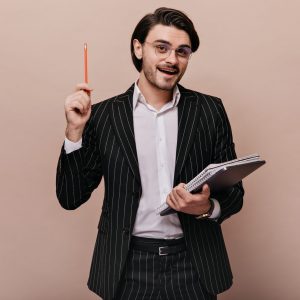 Elegant young teacher with brunette hair, in stylish light shirt, black striped suit, glasses holding writings, pen, and giving lecture. Man standing against beige background